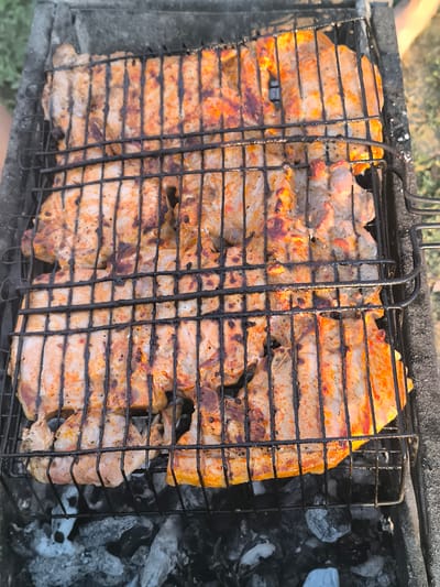 In Ciumai, Moldova, a close-up aerial shot shows pieces of meat, appearing to be pork ribs, cooking on a metal grill basket over glowing embers in a barbecue. The meat is arranged in multiple layers, with grill marks visible on the surface, indicating it is being seared. The background is blurred, suggesting an outdoor setting.