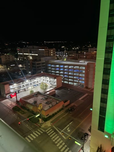 The image presents an elevated nighttime view of downtown Reno, Nevada, United States, looking approximately southwest. The scene is illuminated by numerous artificial light sources against a dark sky.

In the immediate foreground, a light-colored concrete ledge runs along the bottom left. To the right, a segment of a tall, modern building's facade, illuminated vertically with green lights, occupies the right side of the frame.

The midground is dominated by an L-shaped, two-story motel structure, identified by a prominent red "Inn" sign on a pole. This motel, confirmed as the Travelodge Inn, features an internal courtyard parking area containing several parked vehicles and at least two trees. Adjacent to the motel, at a street intersection, is a single-story commercial building with a reddish-brown roof and yellow-tan walls.

Across the street from the motel, a multi-story concrete parking garage is visible, characterized by multiple levels and blue-tinted internal lighting. The street network below includes an intersection with white-striped crosswalks, active traffic lights displaying red signals, and a white left-turn arrow painted on the asphalt. Several vehicles, including sedans and a pickup truck, are visible on the streets and in the motel's courtyard.

The background shows the broader cityscape receding into the distance, marked by scattered lights from various buildings and infrastructure, indicative of an urban environment extending towards a dark horizon where faint distant lights are visible. No individuals are discernible within the frame; the scene depicts static urban infrastructure and vehicles at night.
