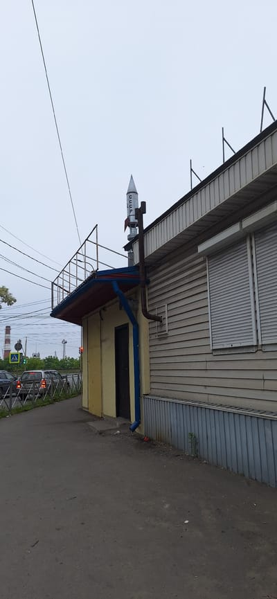 The image captures a street-level view of a building with a rocket-shaped decorative element on its roof. The rocket, painted silver with red fins, has the letters "СССР" vertically inscribed on its side. The building itself appears to be a single-story structure with light-colored horizontal siding and a dark-colored metal base. A dark door is visible on the left side of the building, under a covered awning. A blue downspout runs along the side of the building. The building is situated next to a paved road where several cars are visible. A pedestrian crossing signal with a green light is also present. In the background, industrial structures and trees can be seen under an overcast sky. The overall impression is of an outdoor urban setting.