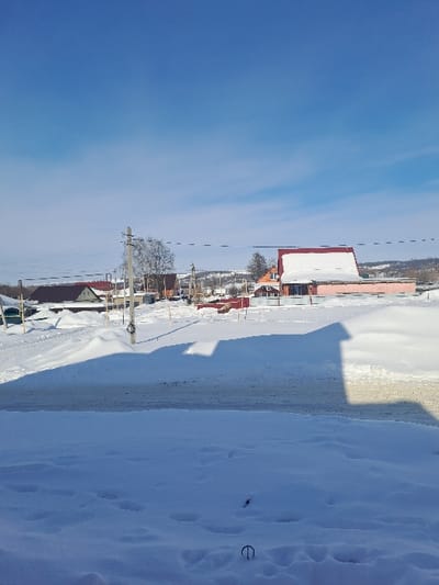 The image displays a snow-covered residential street in Bolshaya Elhovka, Russia, under a clear blue sky with minimal high-altitude clouds. The foreground consists of deep, undisturbed snow, exhibiting a few indistinct indentations and a dark, curved metallic object protruding from the snow near the bottom center. A horizontal strip of dark, partially cleared road separates the immediate foreground from the mid-ground. Long, dark shadows, indicative of structures outside the frame, are cast across the foreground and road, extending from left to right. In the mid-ground, a series of utility poles with multiple overhead wires is visible, along with a bare deciduous tree behind a central pole. Numerous single to two-story residential buildings are present, featuring pitched roofs, many of which are heavily covered in snow. A prominent light pinkish-orange building with a reddish roof and significant snow accumulation is visible on the right side of the frame. The background reveals more scattered structures and distant, snow-dusted hills. No individuals or active subjects are visible in the scene.