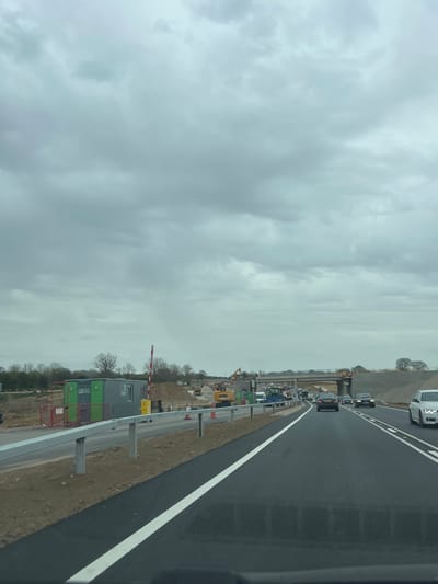 The image shows a road construction site adjacent to a highway. Several vehicles are driving on the highway. Construction equipment, including an excavator, is visible on the construction site. Portable buildings are also present near the construction. The sky is overcast. The location is likely Broadland, United Kingdom, given the context.