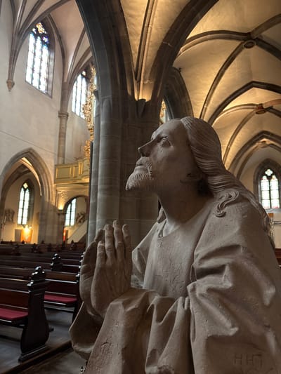 The image shows a stone statue of Jesus Christ in a praying posture, with his hands clasped together and his head tilted upwards. The statue is located inside a church, likely the Saint-Nicholas' Church in Ribeauvillé, France, given the architectural style. The church features high vaulted ceilings, Gothic arches, and stained-glass windows. Rows of wooden pews with red cushions are visible in the background, arranged for congregational seating. The lighting in the church is warm and atmospheric, with light streaming through the windows and illuminating the architectural features. The statue itself appears weathered, with some inscriptions visible on its robe. The overall scene conveys a sense of solemnity and reverence.