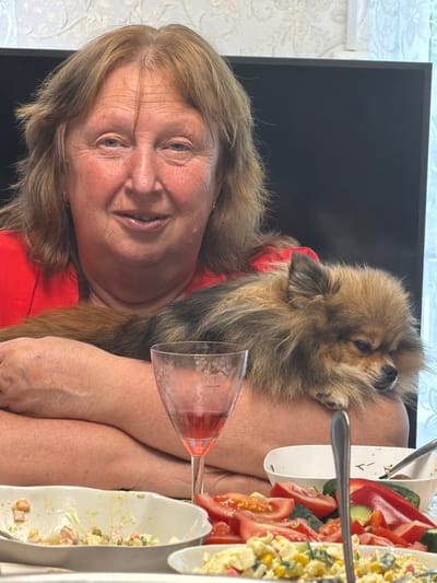 A woman with blonde hair and a red top sits at a table in what appears to be a home environment. She is holding a small, fluffy Pomeranian dog on her lap. The dog is asleep or resting its head on her arm. On the table in front of them are several plates of food, including salads and chopped vegetables like tomatoes and cucumbers. A glass with a pink beverage is also visible. The background includes a dark television screen and a wall with a patterned wallpaper. The lighting suggests it is daytime.
