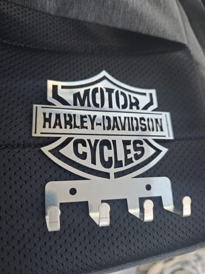 The media file displays a close-up view of a silver Harley-Davidson logo and key holder. The logo is a stylized shield with the words "MOTOR," "HARLEY-DAVIDSON," and "CYCLES" etched into it. Below the logo are four hooks, designed for hanging keys. The key holder appears to be made of metal and is attached to a dark, textured fabric, likely a bag or a piece of clothing. The fabric has a subtle mesh pattern. The lighting suggests an indoor setting or an overcast day, as there are no strong shadows. The image is focused on the product, highlighting its details and design. The scene is static, with no people or specific activity taking place. The location context of Cádiz, Spain, does not provide any specific visual cues within the image itself.