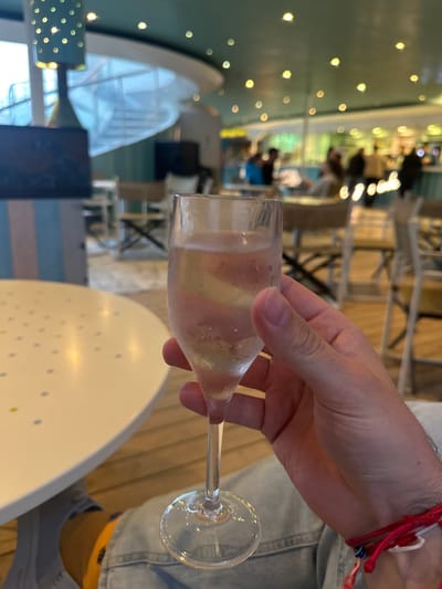 A light-skinned hand, likely male, holding a clear champagne flute containing a pale yellow, effervescent beverage is centered in the foreground. Condensation and bubbles are visible on the glass. The hand has a red string bracelet with a blue bead on the left wrist. The individual is wearing light blue denim pants and a yellow sock is partially visible. To the immediate left is a portion of a white, round table with small yellow and blue dot patterns. The background depicts an indoor establishment identified as being within Puerto del Rosario, Spain, likely a modern bar or lounge. Elements include a striped partition in pastel blue and pink, a green perforated cylindrical light fixture, and a spiral staircase visible through a window or opening on the left. The ceiling is teal with numerous small, warm-toned circular lights. Multiple light-colored tables and chairs are arranged throughout the space. Several blurred individuals are visible in the background, primarily near a dimly lit bar area, suggesting patrons. The floor appears to be a light-colored wood or tile.