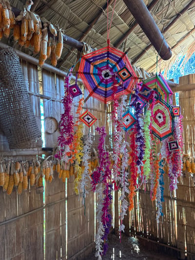 Within a rustic structure, colorful yarn mandalas and diamond-shaped decorations hang from the ceiling and support beams. Corn cobs are strung in rows, adding to the natural ambiance. A woven basket-like object sits below, near a small round object on the wall. The scene evokes a sense of folk art and agricultural traditions, all captured beneath a thatched roof.