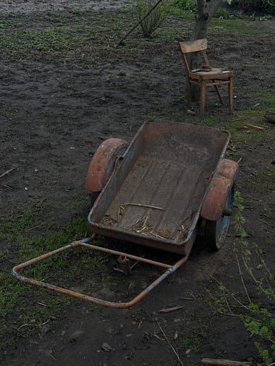 A rusted wheelbarrow sits on muddy ground with sparse grass. A dilapidated wooden chair stands nearby. The location is outdoors, seemingly a yard. No people are visible. The items appear weathered and abandoned. The specific location of Semeno-Aleksandrovka, Russia, provides geographical context.