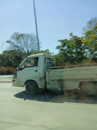 This media file captures a side-view, motion-blurred image of a white flatbed truck driving on a paved road. A man with a beard, wearing a dark t-shirt, is visible driving the truck. The truck has a light-colored flatbed and appears to be carrying some objects, possibly potted plants, in the back.
The scene is set outdoors during the daytime, indicated by the bright blue sky and the shadows cast by the truck. Trees with green foliage are visible in the background, suggesting a temperate climate. The motion blur indicates the photo was taken from a moving vehicle, likely traveling alongside the truck. The overall impression is of a mundane, everyday scene of transportation on a clear day.
There is no visible text in the image. The location context provided is Karatay, Turkey.