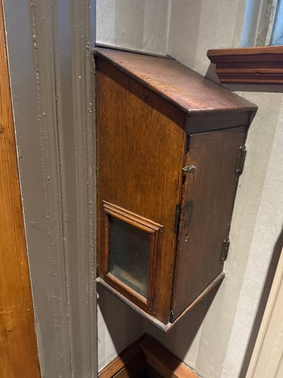 A rectangular, dark-stained wooden box with a hinged lid is mounted above a painted shelf. The box is positioned slightly askew. It features a small, framed glass window on its front-facing side. The lid has two visible hinges. The box sits flush against a horizontally lined wall above the shelf. The shelf is painted a neutral grey-brown color and is partially visible below the box. Wooden trim is also partially visible in the background.