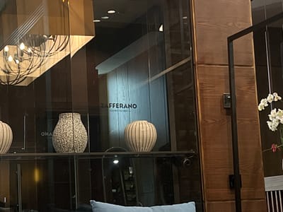 The image depicts an interior scene, likely a restaurant or commercial space, with a prominent display of decorative vases. The focal point is a textured, speckled vase to the left and a ribbed, elongated vase to the right, both situated on a shelf. Behind the vases, a reflective glass surface reveals a sign that reads "ZAFFERANO CUCINA ITALIANA." This suggests the location is likely a restaurant named Zafferano, specializing in Italian cuisine. The background shows a wooden-paneled wall and a chandelier reflected in the glass. A small cluster of white orchids with a pink accent is visible on the far right. The setting appears to be well-lit, with warm ambient lighting.

While the prompt specifies Vyshen'ky, Ukraine, there is no visual information within the image that directly confirms this location. The description is based solely on the visual elements present in the photograph.