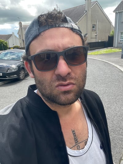 A man is taking a selfie outdoors. He is wearing sunglasses, a grey baseball cap, a black jacket, and a white t-shirt. A tattoo is visible on his chest. In the background, there are houses, a car, and a street. The weather appears cloudy. This scene is consistent with an outdoor setting in the town of Coalisland, United Kingdom.