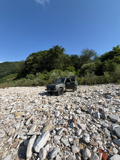 An olive green, four-door off-road vehicle, with its driver's side door and rear hatch open, is parked on a rocky, dry riverbed. The riverbed is densely covered with a variety of light-colored stones, ranging from pebbles to larger, flatter rocks. In the background, a lush, green forest covers the slopes of rolling hills under a clear blue sky. The scene appears to be in a natural, undeveloped area. There is a license plate visible on the vehicle.