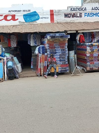The image captures a commercial street scene in Tala ward, Kenya. A young adult male is seated on a yellow cubic object in front of a shop, leaning against a large stack of mattresses. He wears a black baseball cap with white lettering, a red graphic t-shirt, distressed denim jeans, and pink sneakers, with his right hand positioned near his chin. The shop's upper facade, painted white, features a corrugated metal awning. Above the awning, signage reads "AUTHORIZED DEALER MATTRESS" and "KASYOKA HO. MOYALE FASHION" in black and blue text. Underneath the awning, numerous plastic-wrapped mattresses, many displaying colorful patterns and labels such as "RASMI" or "RAIDA," are stacked high on both sides of a dark, open doorway. Various clothing items are hanging on display rails. To the left of the entrance, a blue plastic bucket and other merchandise are visible. The foreground consists of an empty grey asphalt road surface.