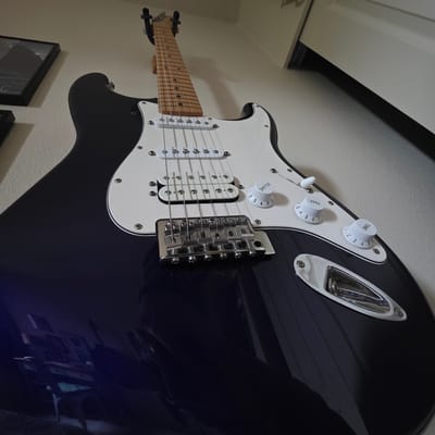 The image shows a close-up, low-angle view of a dark blue electric guitar hanging on a wall. The guitar has a white pickguard, six strings, two white pickup magnets, and chrome hardware. There are three white knobs visible on the pickguard, two labeled "TONE" and one labeled "VOLUME." The guitar is mounted on a wall that appears to be textured and is a neutral, light color. To the left of the guitar, a framed picture is partially visible. The lighting suggests an indoor environment, possibly a room in a home or a music store. The image does not contain any information about the city of Beaverton, United States of America.