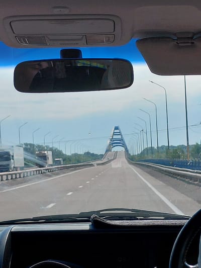 The image shows a view from inside a vehicle, looking forward towards a large arched bridge. The bridge spans across a highway with multiple lanes, indicated by white lines. There are streetlights lining the sides of the highway. In the distance, trees and greenery are visible beyond the highway. There are also some trucks on the highway in the foreground. The sky is partly cloudy. The interior of the car, including the dashboard, rearview mirror, and a sun visor, are visible at the top and bottom of the image. There is no indication of the location being Aqsu city administration, Kazakhstan.