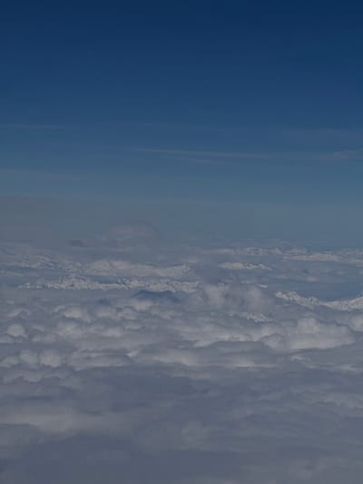 This aerial photograph, captured from an elevated position likely above or in the general vicinity of Oberding, Germany, displays a vast expanse of clouds beneath a clear blue sky, with distant snow-capped mountains visible on the horizon. The lower two-thirds of the frame are filled with a dense, layered blanket of white and grey cumulus and stratocumulus clouds, exhibiting varied textures and internal shadowing. Above the cloud deck, approximately in the upper third of the image, the sky transitions from a lighter blue near the cloud line to a deeper azure at the zenith. In the mid-distance, protruding above the main cloud layer, a range of rugged, snow-covered mountain peaks can be discerned, partially shrouded by atmospheric haze or thinner cloud wisps. No individuals, specific man-made objects, or discernible actions are present in the image. The environment depicted is entirely atmospheric and mountainous.