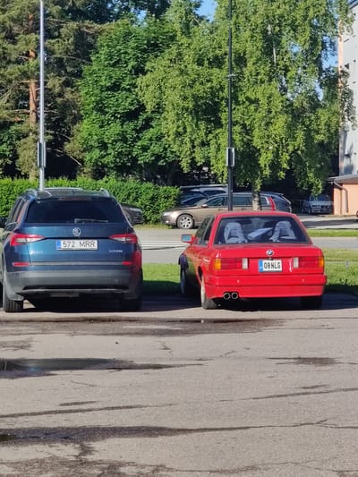 In Saue, Estonia, on a sunny day, two cars are parked parallel to each other in a paved lot. The car on the left is a dark blue Skoda Kodiaq, with its license plate displaying "572 MRR EST". The car on the right is a red BMW, identifiable by its license plate which reads "08 NLG EST". In the background, lush green trees form a dense canopy. A multi-story building with an orange facade is visible to the right of the BMW. The ground is asphalt, with scattered puddles reflecting the bright daylight. A metal pole, likely a lamp post, stands between the two cars and the trees.
