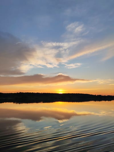 The image depicts a sunset over a body of water, likely a river or lake, near the village of Terekhovo, Russia. The sky is a gradient of blues, oranges, and yellows, with scattered clouds reflecting the warm hues of the setting sun. A dark, silhouetted treeline stretches across the horizon, marking the edge of the land. The water's surface is calm, with gentle ripples distorting the reflection of the sky and the treeline. The overall impression is one of natural beauty and tranquility during twilight.