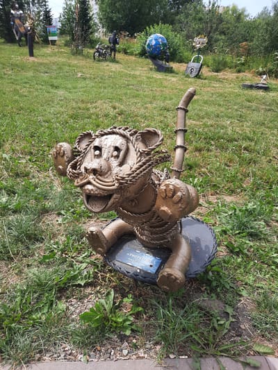 An outdoor metal sculpture of a cartoonish tiger sits on a dark circular base in a grassy area. The tiger sculpture is made of a rough, textured metal, painted a dull brown, and appears to be crafted from twisted ropes and pipes to form its body, limbs, head, and tail. Its eyes are large and hollow, and its mouth is open in a wide grin, revealing a tongue and teeth. It has large paws with visible claws and a long, segmented tail. A small blue plaque with white text is attached to the front of the circular base. The background consists of a moderately cut lawn with scattered weeds and small rocks. In the middle ground and background, other metal sculptures are visible, including a rider on a horse-like creature to the left, a motorcycle with a standing figure to the left-center, a globe on a pedestal, and a large padlock to the right. Trees and buildings are visible in the distance, suggesting an urban or park setting. The sky is overcast. The location is identified as Zelenogorsky, Russia.