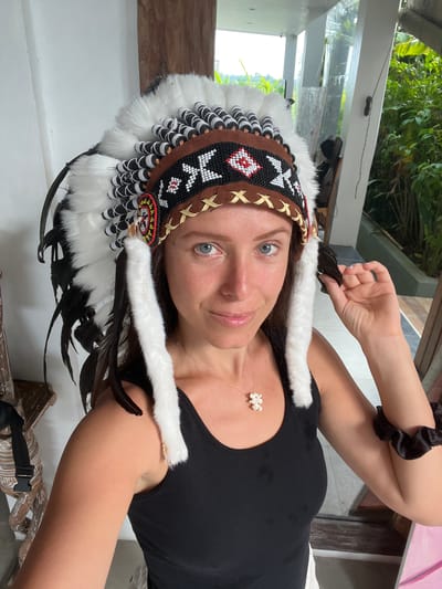 A woman is posing for a selfie indoors, with a Native American-style headdress on her head. She is wearing a black top and a white, flower-shaped necklace. She is fair-skinned with blue eyes. Her hair is long and brown. The location may be Singakerta, Indonesia. A window and some greenery can be seen in the background.
