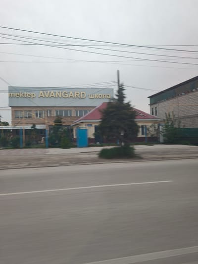 The image depicts a street scene in Qorday, Kazakhstan, featuring a multi-story building identified by a sign reading "mektep AVANGARD школа," which translates to "school AVANGARD." The building has multiple windows. Adjacent to this structure is a smaller building with a red roof. A tree is situated in front of these buildings. The foreground is dominated by a blurred road, suggesting the image was taken from a moving vehicle. Overhead, a series of power lines intersect the sky. A grey sky indicates overcast conditions.