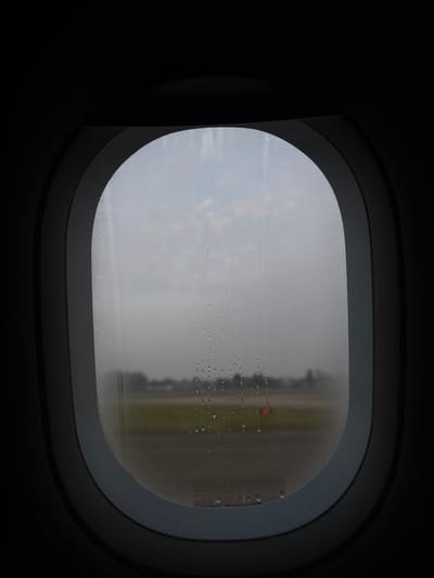 The image shows an airplane window with condensation on the glass. Outside the window, the view is obscured by rain or fog, but glimpses of green grass, a concrete runway, and a red light are visible. The airplane window frame is dark grey and curved, with a layered design. The interior of the airplane around the window is mostly dark. The location in the city of Peschiera Borromeo, Italy is not evident in the image.