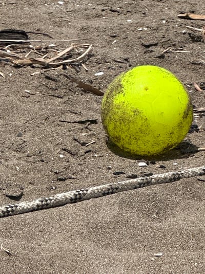 A high-angle shot depicts a yellow ball and a length of rope on a sandy or earthy surface. The ball, positioned in the upper-left quadrant, is somewhat obscured by dark speckling or dirt. A braided rope runs along the right edge of the frame, appearing mostly white and grey. The surface is textured with debris. The location is implied by the prompt, but no specific evidence in the image confirms it is Ortaca, Turkey.