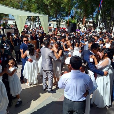 The image shows a mass wedding event occurring in Cerro Colorado, Peru. A large crowd of people, many dressed in wedding attire, are gathered outdoors on a paved area, likely a plaza. Numerous couples are seen kissing, suggesting a shared wedding ceremony. The brides are wearing white dresses, while the grooms are in suits of various colors, including gray and blue. Spectators are present, some holding up phones, possibly to record the event. A stage or elevated area is visible in the background, with sound equipment, including speakers. A white tent or canopy offers shade. The Peruvian flag is seen flying in the background. Trees surround the area, indicating an outdoor setting. The event seems to be of a significant scale, involving a substantial number of participants and attendees.