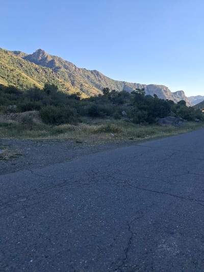 This media file captures a scenic view of a mountainous landscape in Shatin, Armenia. The scene is dominated by a steep, verdant mountainside on the left, covered in dense foliage and trees. The mountains are rugged and show signs of geological formations, with rocky outcrops visible. A clear, bright blue sky fills the upper portion of the image, suggesting a sunny day.
Adjacent to the mountainside, a dark, paved road stretches diagonally across the frame, taking up the right half of the image. The road appears to be asphalt and shows some signs of wear, with visible cracks. The perspective suggests the viewer is either on or very near the road, looking up towards the mountains.
The time of day appears to be morning or early afternoon, given the bright sunlight casting shadows on the mountainside and illuminating the vegetation. The weather seems clear and pleasant. There are no people or specific objects of interest on the road or in the immediate landscape, focusing the attention on the natural environment. The overall impression is one of a remote and natural setting, likely a roadside view in a mountainous region. No text is visible in the image.