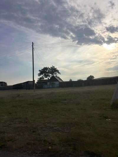 The media file depicts an outdoor scene, oriented vertically. The sky dominates the upper portion, filled with dramatic clouds ranging from dark gray to wispy white, with the sun partially obscured, suggesting late afternoon or early morning. Power lines stretch across the frame, supported by a wooden pole.
Below the sky, a wooden fence runs horizontally, separating a grassy field from what appears to be a rural or historical settlement. A lone tree grows near the fence, its branches reaching towards a small, rustic building with decorative patterns on its side. More structures are visible behind this building, some appearing weathered. The ground is covered in dry grass, with hints of darker soil. The overall atmosphere is somewhat somber due to the cloudy sky, but the presence of the sun hints at potential warmth. There is no visible text in the image. The setting appears to be a rural area in Novonikolayevka, Russia.