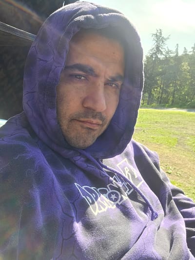 A man is wearing a purple hoodie with white lettering and a hood. The hoodie has a crackle pattern. The background shows green grass and trees illuminated by sunlight.
The location is not discernible from the image. There is no evidence to indicate that this image was taken in Chargan, Bulgaria.