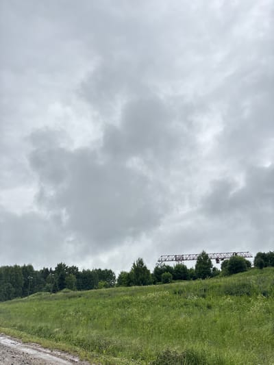 The image shows a cloudy sky over a grassy hillside in Mendeleevo, Russia. A large, red industrial gantry crane is visible in the distance, silhouetted against the overcast sky, positioned above a dense line of trees. The foreground features a dirt road with some puddles, suggesting recent rain. The hillside itself is covered in lush green grass and wildflowers, with scattered trees and bushes. The overall impression is a somewhat bleak, rural landscape under a heavily clouded sky.