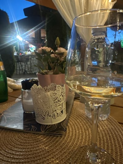 A close-up, low-angle, vertical shot shows a wine glass filled with white wine on a wooden table. The glass has a reflection of a blue sky and a tent-like structure on its surface. In the background, a salt shaker with a black lid and a white doily-like holder are visible. A green bottle is on the left, and a potted plant with pink flowers is behind the salt shaker. The scene appears to be outdoors, possibly at a restaurant or bar in Dragoman, Bulgaria. The lighting suggests it is dusk or early evening.