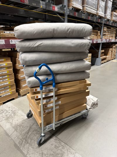 The image depicts the interior of a large warehouse, possibly a retail store, located in Kaiserslautern, Germany. Racks of stacked cardboard boxes extend into the background, while a flatbed trolley loaded with goods is in the foreground. The trolley is filled with gray cylindrical items, possibly packaged bedding, and stacks of long, rectangular cardboard boxes. The trolley has a blue handle and silver frame, with small wheels at the corners. The environment suggests a storage or distribution area, with a focus on inventory and logistics.