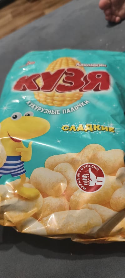 A bag of "Kuzya" brand sweet corn puffs is displayed on a dark gray surface, possibly a couch or blanket. The bag features a bright turquoise background with a large, stylized image of a corn cob and the brand name "KUZYA" in bold red lettering. Below the brand name, the text "КУКУРУЗНЫЕ ПАЛОЧКИ" (corn puffs) is printed in white. A yellow cartoon character, resembling a dinosaur with big eyes and wearing a blue and white striped shirt, is positioned on the left side of the bag, pointing with its finger. To the right, the word "СЛАДКИЕ" (sweet) is written in yellow letters. A circular red and white seal with a thumbs-up icon and the text "МАРКА № 1 В РОССИИ" (Brand No. 1 in Russia) is visible on the right side of the bag, overlaying images of the corn puffs themselves. The lighting suggests an indoor setting, with the bag illuminated from above. There is no information in the image that places it in Cherepovets, Russia.
