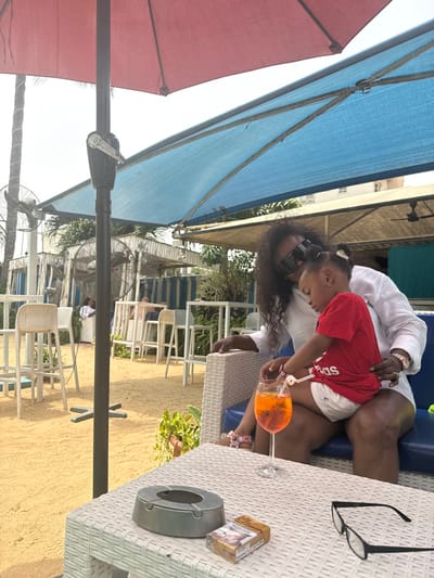 The image captures an outdoor, sunny scene, likely at a resort, beach club, or poolside cafe. The primary subjects are an adult, presumably female, and a young child, who appears to be sitting on the adult's lap or very close to her at a table. The ground is a light, sandy color.

The adult has long, dark, curly hair with sunglasses pushed up onto her head. She is wearing a white, long-sleeved top and light-colored shorts, and a watch on her left wrist. The child, with dark hair and a hair accessory, is wearing a red t-shirt with a white logo (partially visible, resembling "ADIDAS") and white shorts.

They are seated at a white, woven-texture outdoor table. On the table in front of them, there is a clear stemmed glass containing an orange-colored drink, resembling an Aperol Spritz. The adult is holding this glass, and the child is looking intently at it. Another clear plastic novelty cup with a straw, also containing an orange drink, is visible next to the child. Further to the right on the table, a pair of black-framed eyeglasses, a silver-colored ashtray, and a pack of "Marlboro" cigarettes in gold and orange packaging are visible. There is also a small, gold-framed picture on the table.

The scene is shaded by large sun umbrellas. A prominent blue umbrella is overhead, extending across the left side of the frame, and a smaller red umbrella is partially visible further left. The bright sky is visible between the umbrellas. In the background, there are other white outdoor tables and chairs, typical of a cafe setting. Electric fans are visible, some attached to umbrella poles and others standing on the ground, suggesting warm weather. Beyond the seating area, there are some buildings and greenery. The overall atmosphere appears relaxed and casual, characteristic of a vacation or leisure activity.