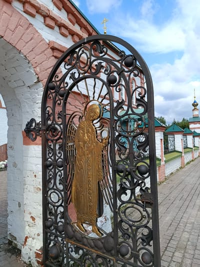 A full shot from a low angle shows a closed, black wrought-iron gate with a golden angelic figure in the center. The gate is ornate, with swirling metalwork and spherical accents. Behind the gate, a white brick wall with pink brick accents forms an arched entryway to a monastery. The monastery's red and white facade is visible in the background, along with a turquoise-domed cupola. A cobblestone path leads away from the gate toward the monastery. The sky is a bright blue with scattered white clouds. The overall scene depicts a religious or historical site.