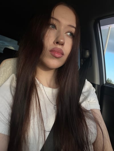 A young woman with long, dark brown hair is in the passenger seat of a car, looking out the window. She is wearing a white t-shirt and a silver necklace with a charm. The sun is shining, casting shadows in the car. The background shows a cloudy sky and some trees. The location is the city of Gavrilovka, Russia.