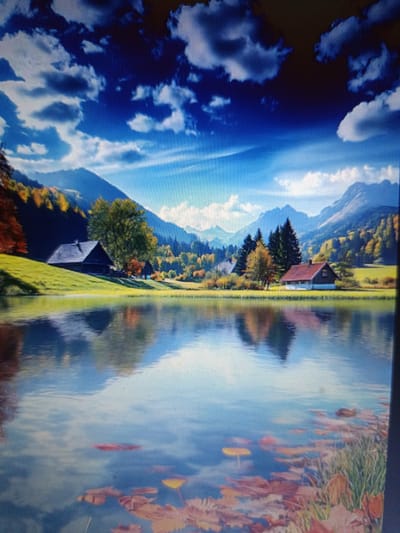 The image depicts a serene alpine landscape during autumn. In the foreground, a calm body of water reflects the sky and surrounding scenery. Scattered on the water's surface are fallen leaves, some in shades of red and yellow. Beyond the water, a gently sloping grassy hill leads up to a cluster of buildings.
On the left side of the hill, a dark-colored, rustic house with a steeply pitched roof sits nestled amongst trees displaying vibrant autumn foliage. Further to the right, a second building with a reddish-brown roof is visible, set against a backdrop of dense evergreen trees and mountains. The mountains in the distance are tinged with the warm colors of autumn, with patches of green and yellow from trees. The sky above is a brilliant blue, punctuated by dramatic, dark and light-colored clouds that create a sense of depth and movement.
The scene evokes a peaceful, natural environment, possibly a rural or semi-rural setting. It is not possible to determine the specific location within the city of Košice, Slovakia, as the image portrays a generic mountain landscape that could be found in various regions with similar topography and climate. There are no people or animals visible in the image, and the focus is on the natural beauty and the quaint structures.