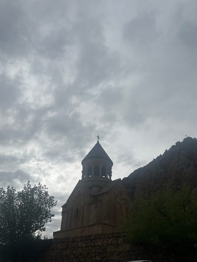 This image depicts a historical stone church, identified as located in the city of Amaghu, Armenia, framed against an overcast sky and rugged terrain. The church occupies the central midground, featuring a distinct domed cupola surmounted by a cross at its apex. The structure is constructed from light-colored stone, appearing somewhat darkened by shadow, with visible architectural details on its facade. To the immediate right and extending into the background, a steep, dark mountainous or rocky formation rises sharply. A small flag or marker is discernible near the peak of this formation on the far right. The left foreground and midground are partially occupied by a dark, leafy tree, obscuring the lower portion of the church and a stone wall. This stone wall, composed of irregular, rough-hewn stones, stretches across the lower section of the image, forming a base for the structures and vegetation. The sky is entirely covered by dense, grey clouds, creating a diffused and somewhat somber illumination across the scene. No individuals or active interactions are present.