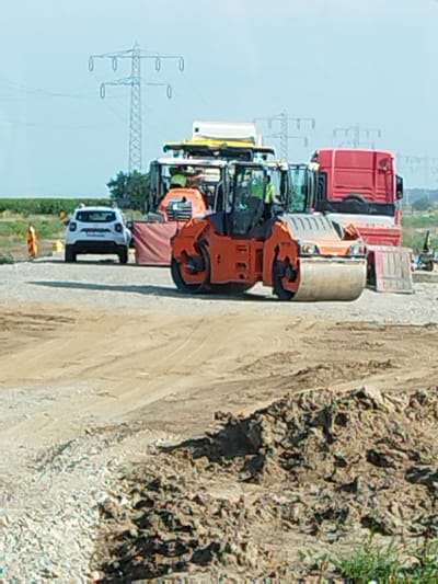 This image depicts a construction site with heavy machinery in Nucet, Romania. A large orange steamroller is in the foreground, facing right, with its large cylindrical drum visible. Behind the steamroller, a white passenger car is parked. Further in the background, a red semi-truck is parked, and various construction vehicles, including what appears to be a front-end loader or a similar piece of equipment with a visible operator wearing a yellow vest, are present. Power transmission towers and lines are visible against a clear sky in the distance, suggesting an open, possibly rural or exurban, environment. The ground is a mix of dirt, gravel, and possibly some debris, indicative of ongoing construction or roadwork.