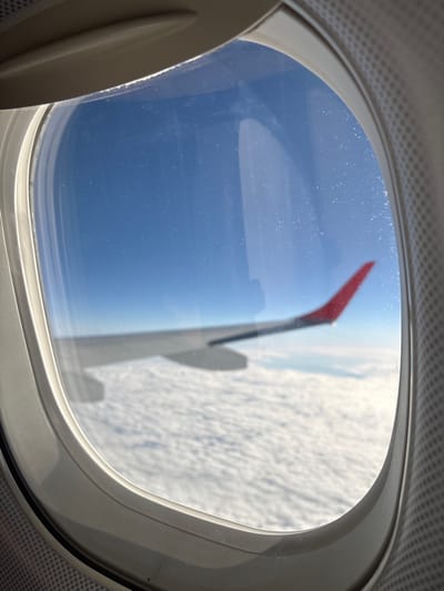 The image depicts the view from an airplane window. A portion of the airplane's wing is visible, with a red winglet. The sky is blue above, and below the wing is a white, cloud-like expanse. The window is oval-shaped and surrounded by interior paneling of the aircraft. Considering the winglet design and the location of Schwechat, Austria, it is highly likely that the aircraft is operated by Austrian Airlines, as Schwechat is the location of Vienna International Airport, their hub.