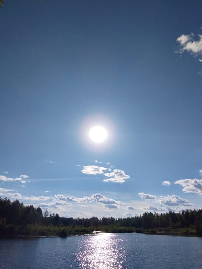 The image depicts a serene natural landscape under a bright, sunny sky. The main subjects are a calm body of water in the foreground and a dense line of trees forming a forest along the far bank. The sky is a clear, vibrant blue, populated by scattered white clouds. A prominent feature is the sun, shining intensely in the upper center of the frame, casting a bright glare and creating a sparkling reflection on the water's surface. The sun's brightness suggests it is midday.
The scene is a natural setting, likely a lake or a wide river, bordered by a lush forest of predominantly dark green coniferous trees, with some deciduous trees and undergrowth nearer the water's edge. The water is dark blue and exhibits ripples, indicating a gentle breeze. The reflection of the sun on the water is particularly striking, appearing as a path of shimmering light.
The overall mood conveyed by the image is peaceful and tranquil. The clear sky and bright sunlight suggest good weather, likely during the warmer months. The location cues, such as the type of forest and the overall landscape, align with a setting in Russia, possibly near the "Go Maya" region. There are no people or man-made objects visible, emphasizing the untouched natural beauty of the scene. The image captures a moment of natural splendor, highlighting the interplay of sunlight, water, and vegetation. No text is visible in the image.