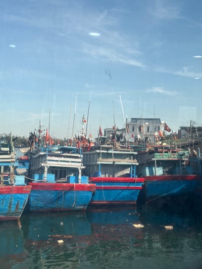 This image depicts a fleet of brightly colored fishing boats docked in a harbor. The boats are predominantly blue and red, with some featuring green accents. They are equipped with numerous poles and masts, suggesting their readiness for fishing. Red flags are prominently displayed on many of the vessels. In the background, a large white building with a grey roof and several windows is visible, along with other structures and trees. The water in the harbor is murky and contains some debris. The sky above is clear and blue with some wispy white clouds. This scene is likely located in the city of An Hải, Vietnam, a region known for its vibrant fishing industry and coastal landscape.