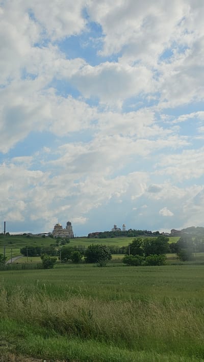 The image depicts a pastoral scene with a wide expanse of green grass in the foreground, stretching towards a distant hillside dotted with trees and buildings. The sky is a prominent feature, filled with fluffy white clouds against a backdrop of pale blue.

The scene appears to be set in Yessentuki, Russia, given the location context. The buildings on the hillside, particularly the one with golden domes, suggest a possible religious or historical site. The overall atmosphere is serene and peaceful, indicative of a pleasant day, likely in the late morning or early afternoon, judging by the bright, diffused light. The weather seems fair, with the clouds suggesting a partly sunny day. There are no visible people or specific activities taking place, making it a landscape-focused image. The tall grass in the foreground is dry and wispy, hinting at warmer weather, possibly late spring or summer. The composition emphasizes the vastness of the landscape, with the sky dominating the upper portion and the rolling green fields leading the eye towards the distant structures.
