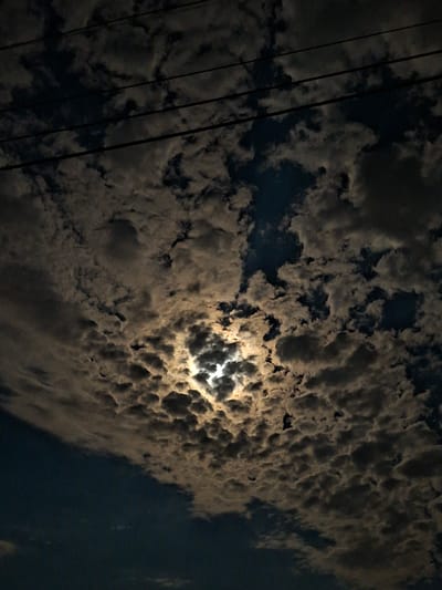 The image depicts a night sky filled with clouds in Galabovo, Bulgaria. A bright light, possibly the moon, shines through a break in the cloud cover, illuminating the surrounding clouds with a golden glow. Electrical power lines are visible in the left portion of the frame.