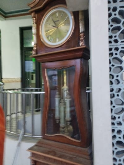 A slightly blurry, eye-level, full shot captures a tall, ornate grandfather clock situated indoors in Kecamatan Panji, Indonesia. The clock, made of polished dark brown wood, is the central subject, positioned towards the right of the frame. Its upper section features a large, circular clock face with a golden metallic finish and black Roman numerals, indicating a time of approximately 10:10 with golden hands. The clock's crown and sides are adorned with intricate wooden carvings.
Below the clock face, a glass panel reveals the clock's inner mechanisms, specifically three metallic pendulum weights: two elongated, tubular weights and a larger disc-shaped pendulum bob at the bottom. The wooden casing around this glass section has a graceful, curved design. The clock's base features tiered wooden detailing.
To the left of the clock, a metallic railing suggests an indoor balcony or corridor. Beyond the railing, a dark door or window with a grid pattern is visible. The wall in the background on the left is white with a thin green and white trim near the ceiling. A fragment of an orange garment, possibly belonging to a person, is visible on the far left edge of the image. On the far right, a white architectural pillar or wall stands beside a decorative grey screen or lattice, featuring an intricate, repeating geometric pattern. The scene is static, and no specific activity or event is taking place. The lighting suggests it is daytime. No legible text is visible anywhere in the image.