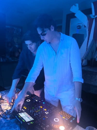 Two men are operating a DJ setup in a dimly lit room, likely at a party or event, in Pozuelo de Alarcón, Spain. The man in the foreground, wearing a light blue collared shirt and white pants, is actively adjusting controls on the DJ console, with his hands positioned over knobs and buttons. He is also wearing sunglasses and a wristwatch. The man behind him, dressed in a dark blue shirt, also appears engaged with the equipment, his hands hovering near the console. The DJ setup, a "Pioneer Dj" unit, features illuminated buttons and screens. The overall ambiance is enhanced by blue lighting, which casts a cool tone over the scene. The background is blurred but suggests a room with some decorative elements and possibly a mirror. The presence of a patterned tablecloth underneath the DJ equipment indicates a domestic or semi-private setting.
