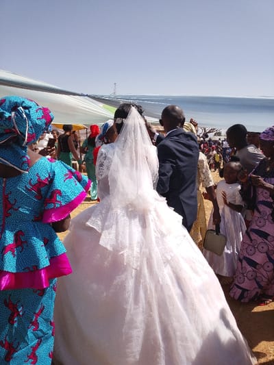 An outdoor wedding scene is depicted in Angwan Sarki, Nigeria, under a clear, bright sky. The central figure, a bride, is seen from behind, wearing a white, voluminous wedding gown with a sheer, sparkling overlay and a long white veil secured by a sparkling tiara. To her right, a groom in a dark suit adjusts something near her head.
To the immediate left of the bride, another woman wears a vibrant blue and pink patterned top with a horse motif, and a matching headwrap. She holds a smartphone vertically in her left hand, appearing to record or photograph the event. Further left and in the background, other individuals in various traditional and modern attire are visible, some holding phones. One woman in the mid-ground wears a red headscarf and a green garment.
To the right of the groom, a young girl in a white dress holds a small, rectangular grey bag. Behind her, an adult woman in a patterned purple dress with circular motifs looks towards the front. A crowd of additional guests is visible in the background, partially obscured, suggesting a large gathering.
The ground is dry and appears to be earth or sand. Overhead, a large white and light blue fabric canopy, possibly a tent or awning, is stretched across the scene, providing shade. In the distant background, a tall, slender metallic structure, possibly a communication tower, is visible against the clear sky.