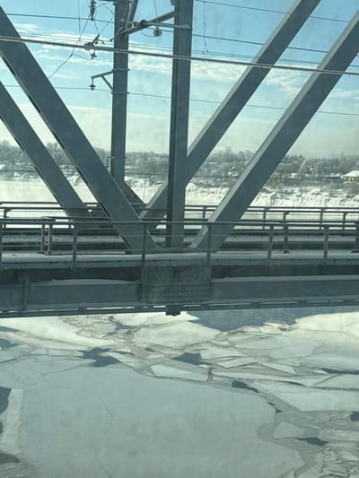 The image displays a forensic view of the Volkhov River Railway Bridge in Volkhov, Russia, during winter. The scene is dominated by the bridge's grey metal truss structure, composed of robust diagonal and vertical beams with visible bolted or riveted connections. Overhead, railway electrification lines, including catenary wires and insulators, are discernible against a partly cloudy, bright blue sky.

Below the bridge deck, the Volkhov River is largely covered by an expanse of fragmented ice floes, interspersed with patches of dark open water, indicating freezing conditions. A metal walkway or service platform runs along the bridge, equipped with railings and partially covered in snow.

The distant riverbank and landscape are blanketed in snow, featuring numerous bare deciduous trees and scattered buildings, including a light-colored structure on the right side of the far bank. The perspective is from an elevated position, likely from a train or vehicle traversing the bridge, with reflections visible on the viewing surface. No individuals or specific actions are depicted within the frame.